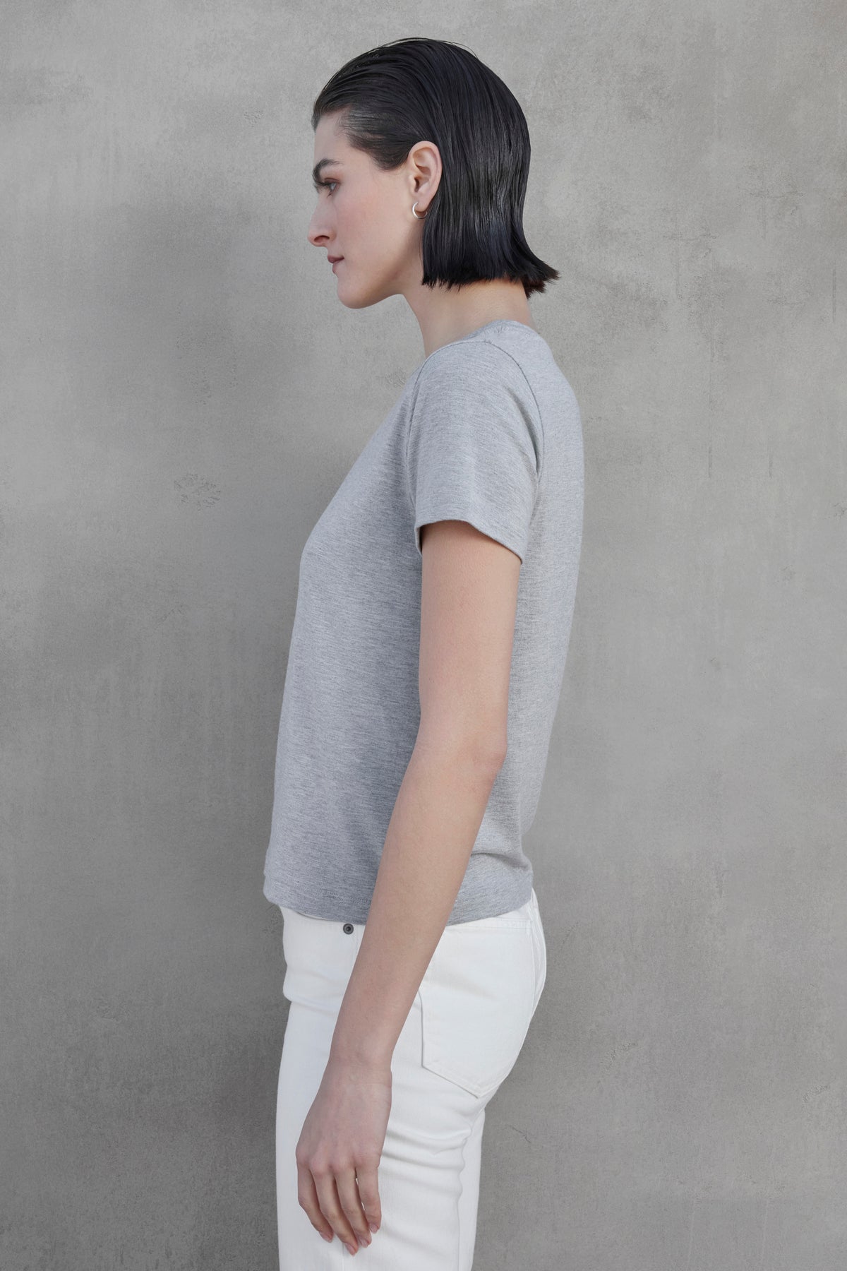   Side profile of a person with short dark hair, wearing the SOLANA TEE by Velvet by Jenny Graham and white pants with shrunken sleeves, standing against a grey textured background. 