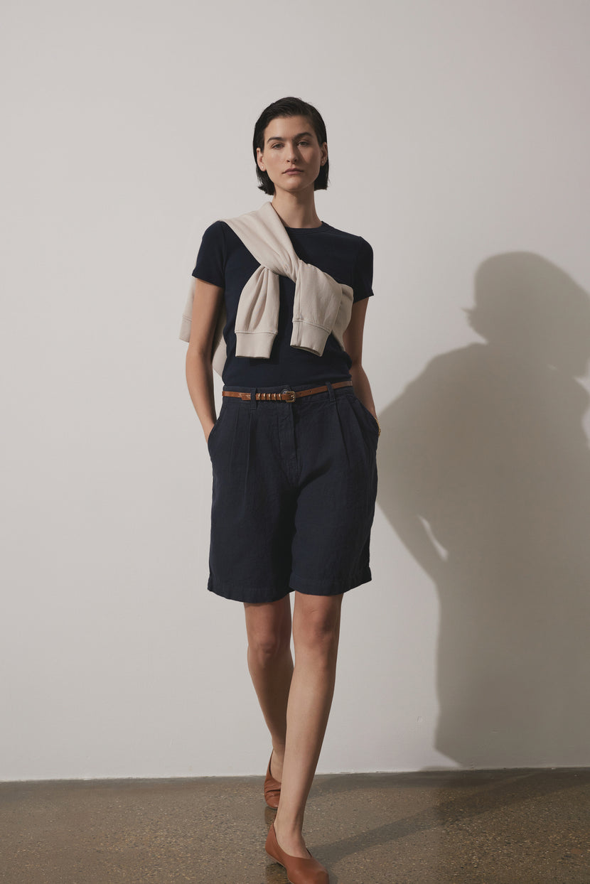 A woman in a stylish outfit with LARCHMONT HEAVY LINEN SHORTS by Velvet by Jenny Graham, cream blouse, and tan belt poses against a plain wall, casting a shadow.