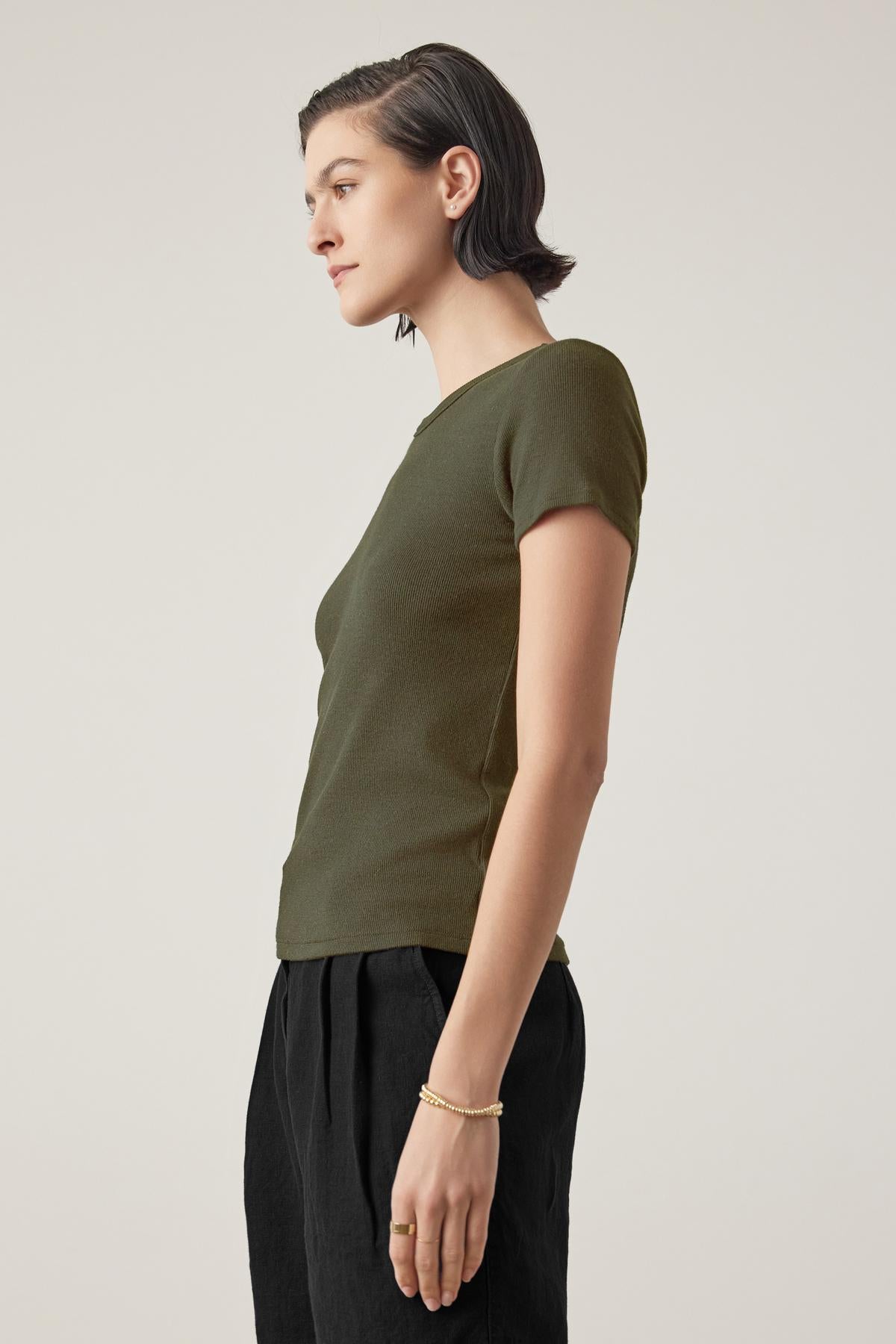   Side profile of a person with short dark hair, wearing an olive green BEDFORD TEE by Velvet by Jenny Graham with a fitted silhouette and black pants, standing against a plain background. 