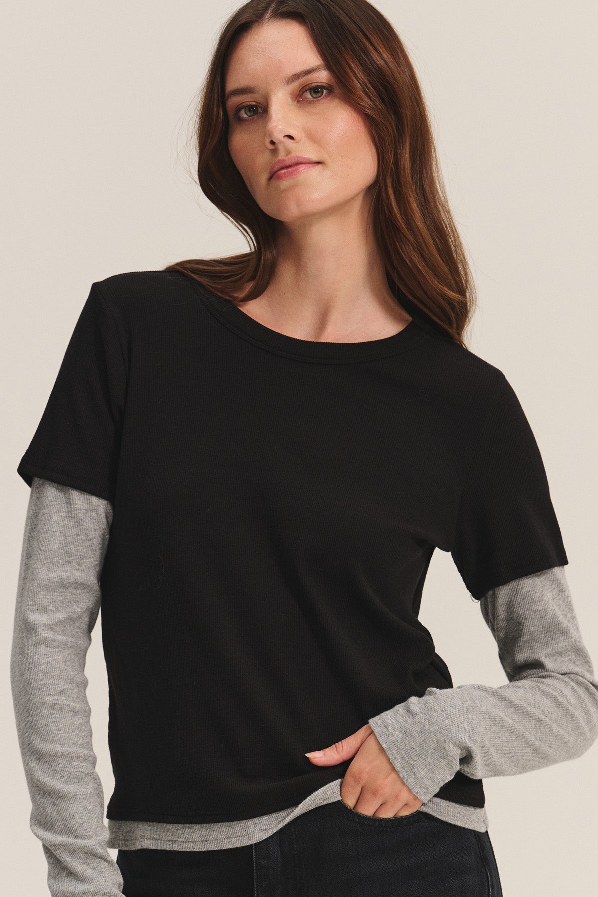 A woman with long brown hair wears the Velvet by Graham & Spencer BRONSON TEE, a black modal rib crewneck, layered over a gray long-sleeve shirt for a chic double-layered look against a simple background.-40681598681281