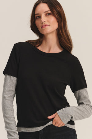 A woman with long brown hair wears the Velvet by Graham & Spencer BRONSON TEE, a black modal rib crewneck, layered over a gray long-sleeve shirt for a chic double-layered look against a simple background.