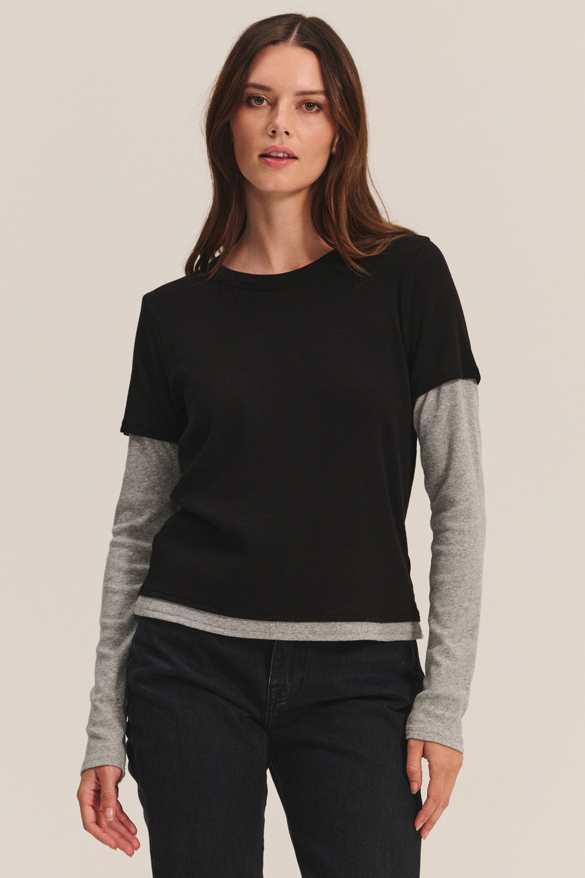 A woman stands against a plain backdrop wearing the Velvet by Graham & Spencer BRONSON TEE in black, layered over a gray long-sleeve shirt and paired with dark pants for a sleek, modern double-layered style.-40681598648513