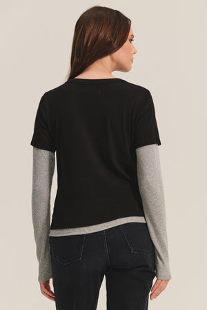 A person with long brown hair is seen from the back, wearing the Velvet by Graham & Spencer BRONSON TEE—a black modal rib crew neck—layered over a gray long-sleeve shirt and paired with dark jeans for a stylish, double-layered look.