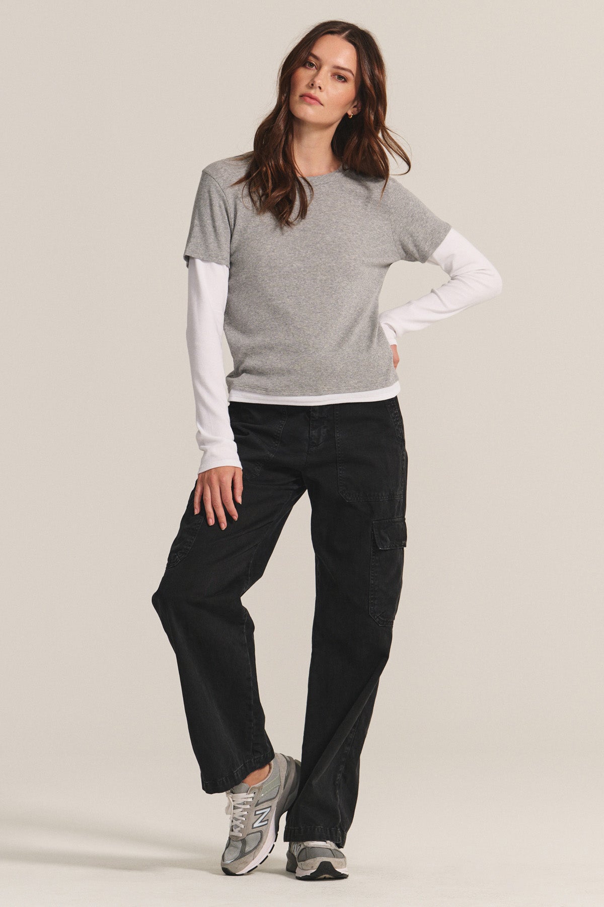 A woman stands against a plain background, wearing the BRONSON TEE by Velvet by Graham & Spencer—a gray ribbed crewneck styled over a white long-sleeve shirt—paired with black cargo pants and gray sneakers.-40334122320065