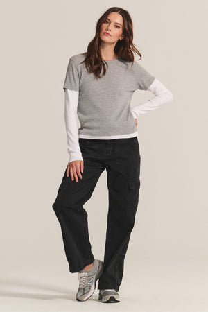 A woman stands against a plain background, wearing the BRONSON TEE by Velvet by Graham & Spencer—a gray ribbed crewneck styled over a white long-sleeve shirt—paired with black cargo pants and gray sneakers.