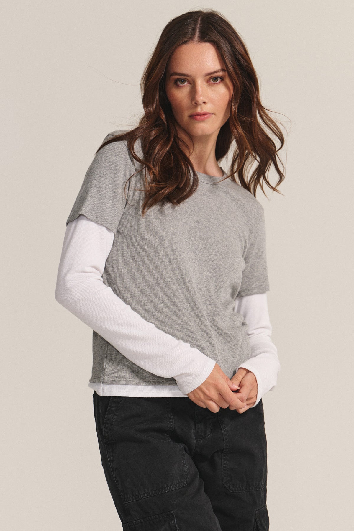 A woman stands against a plain background, wearing the BRONSON TEE by Velvet by Graham & Spencer over a white long-sleeve shirt and black pants, showcasing a double-layered style while looking directly at the camera.