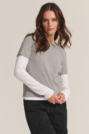 A woman stands against a plain background, wearing the BRONSON TEE by Velvet by Graham & Spencer over a white long-sleeve shirt and black pants, showcasing a double-layered style while looking directly at the camera.