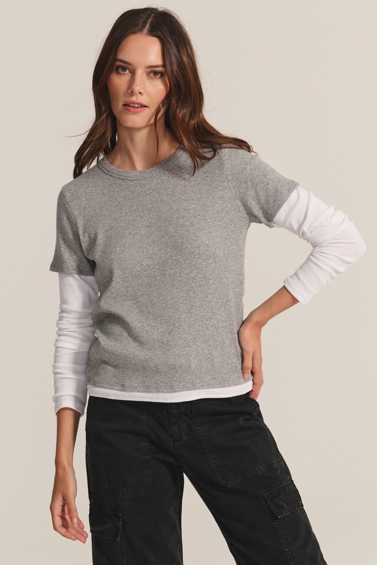 A woman poses against a plain background, layering the BRONSON TEE by Velvet by Graham & Spencer—a grey modal rib crewneck—over a white long-sleeve shirt with black cargo pants.