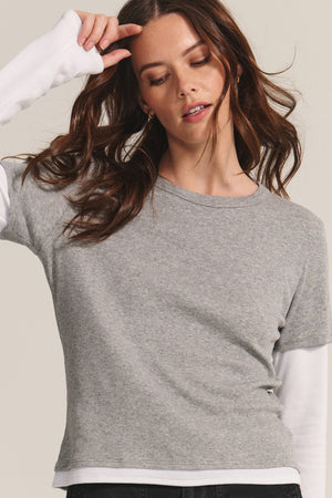 A woman wears the BRONSON TEE by Velvet by Graham & Spencer, layering the grey crewneck over a long-sleeve white shirt, eyes closed and hand raised to showcase a chic, modern double-layered style.