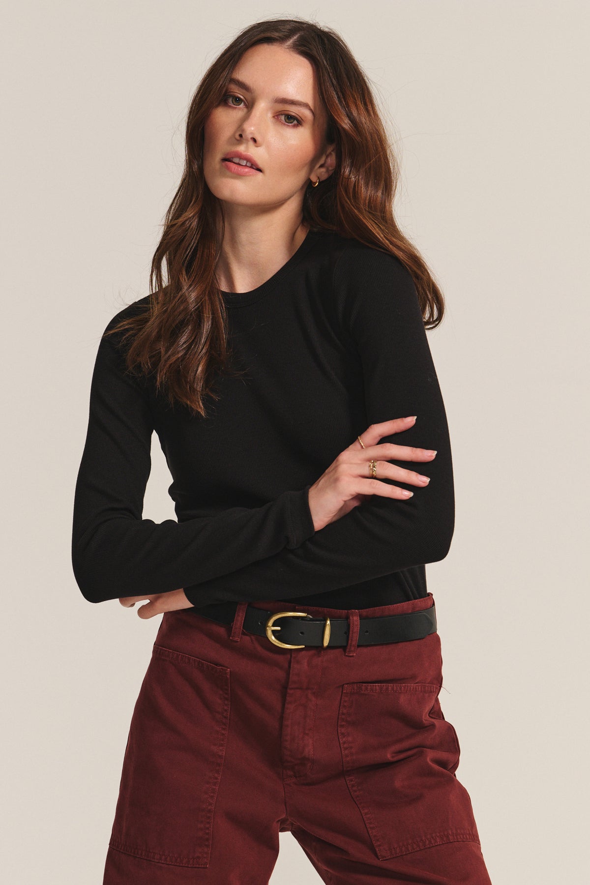   A woman with long brown hair wears the CAMINO TEE by Velvet by Jenny Graham, in black modal rib, paired with burgundy pocketed pants and a black belt with a gold buckle, posed against a plain background. 