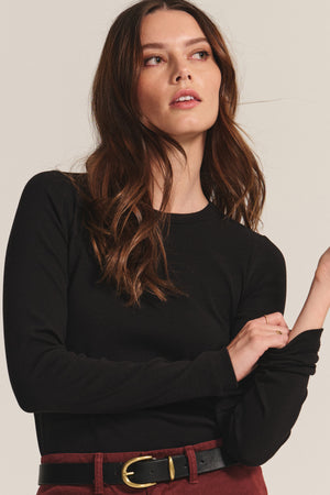 A woman with long brown hair wears the Velvet by Jenny Graham CAMINO TEE in black, paired with maroon pants and a black belt, posing against a neutral background.
