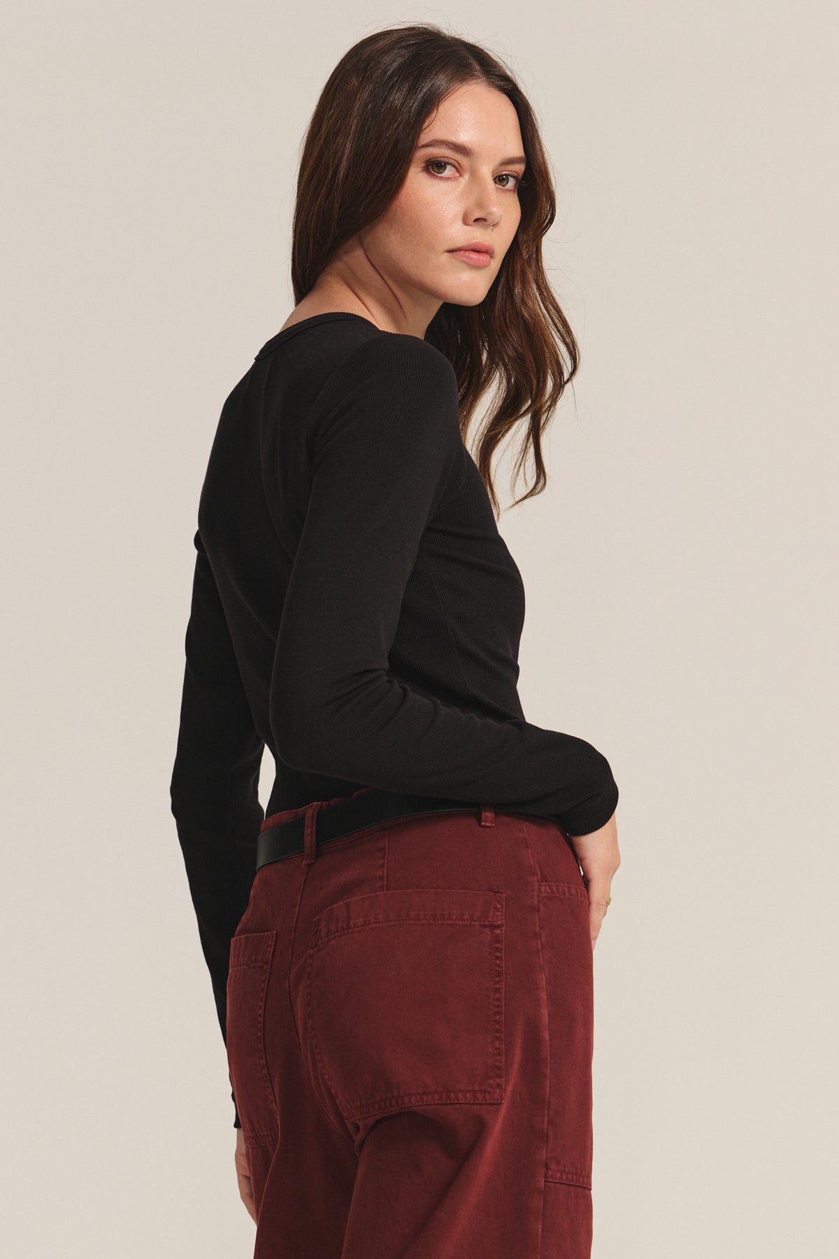   Woman with long brown hair wears the CAMINO TEE by Velvet by Jenny Graham in black, paired with high-waisted maroon pants, standing and looking over her shoulder against a light background for a slimming look. 