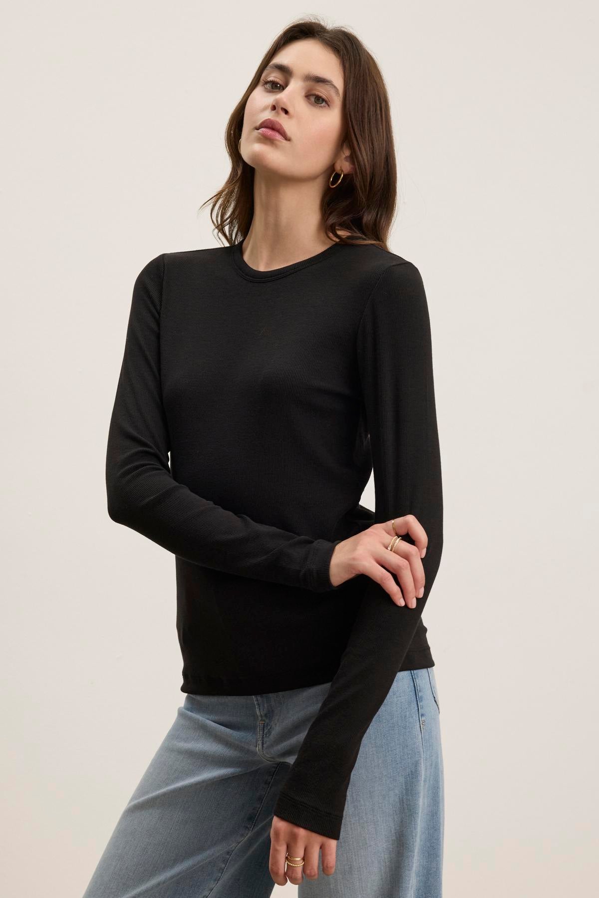 A person wearing the CAMINO TEE by Velvet by Jenny Graham, a black long-sleeve shirt crafted from soft modal rib fabric, pairs it with blue jeans against a plain background. This outfit highlights the top's slimmer fit, making it perfect for versatile layering.-38352888889537