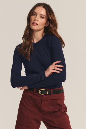 A woman with long brown hair wears the CAMINO TEE by Velvet by Jenny Graham, paired with maroon high-waisted pants and a black belt, standing with arms crossed against a neutral background.