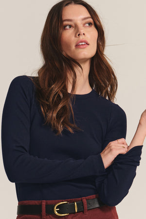 A woman with long brown hair wears the Velvet by Jenny Graham CAMINO TEE in navy, a slim fit modal rib layering top, styled with a brown belt featuring a gold buckle. She looks slightly upward and to the side.