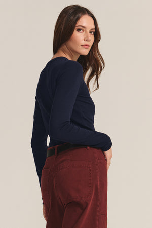 A woman with long brown hair models the Velvet by Jenny Graham CAMINO TEE, a navy slim-fit modal rib long-sleeve layering top, paired with maroon pants as she stands slightly turned, looking over her shoulder.