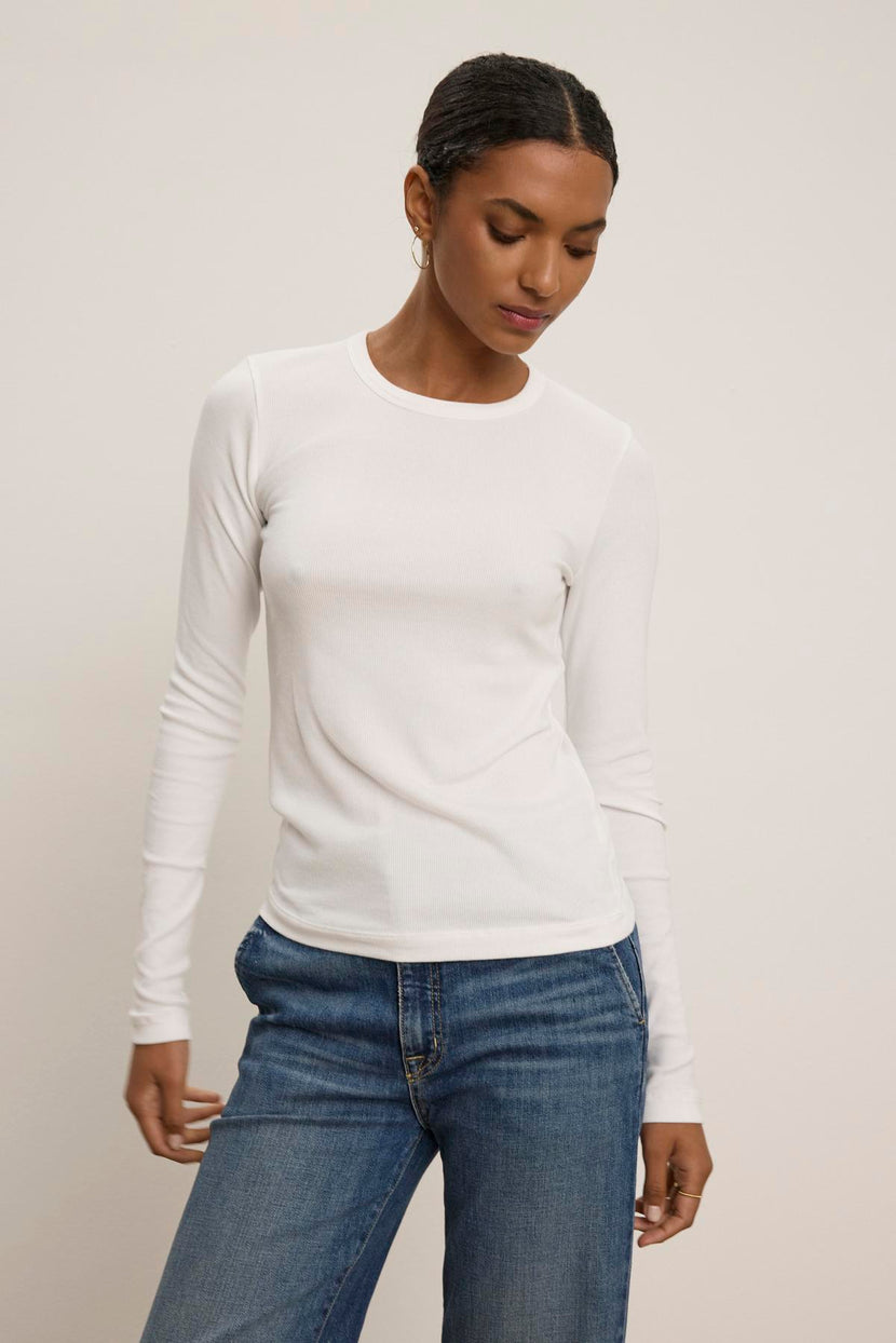 Clad in the elegant CAMINO TEE by Velvet by Jenny Graham paired with slimmer-fit blue jeans, a person stands against a plain background, showcasing a versatile style.