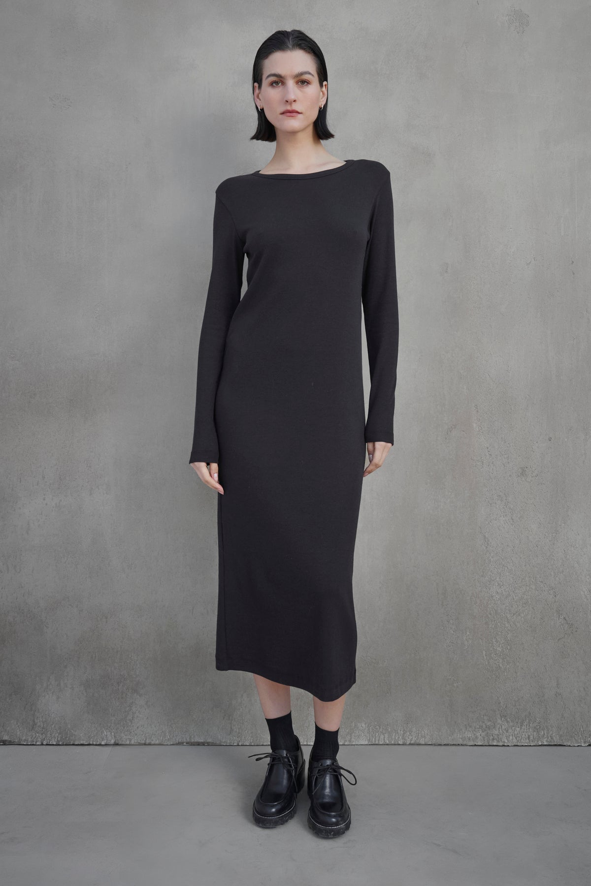   A person stands against a plain concrete wall, wearing the CORONADO DRESS by Velvet by Jenny Graham, which is a long-sleeved, ankle-length black dress, paired with black shoes. 