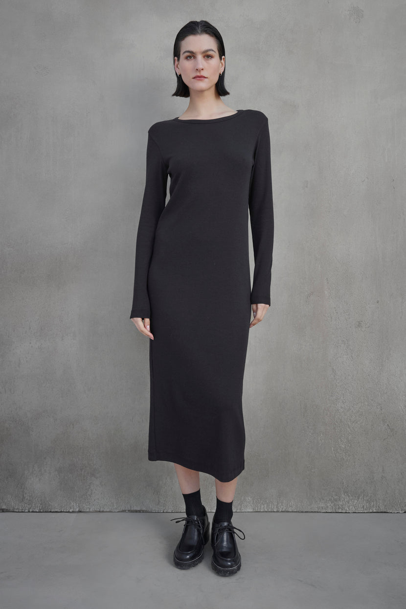 A person stands against a plain concrete wall, wearing the CORONADO DRESS by Velvet by Jenny Graham, which is a long-sleeved, ankle-length black dress, paired with black shoes.