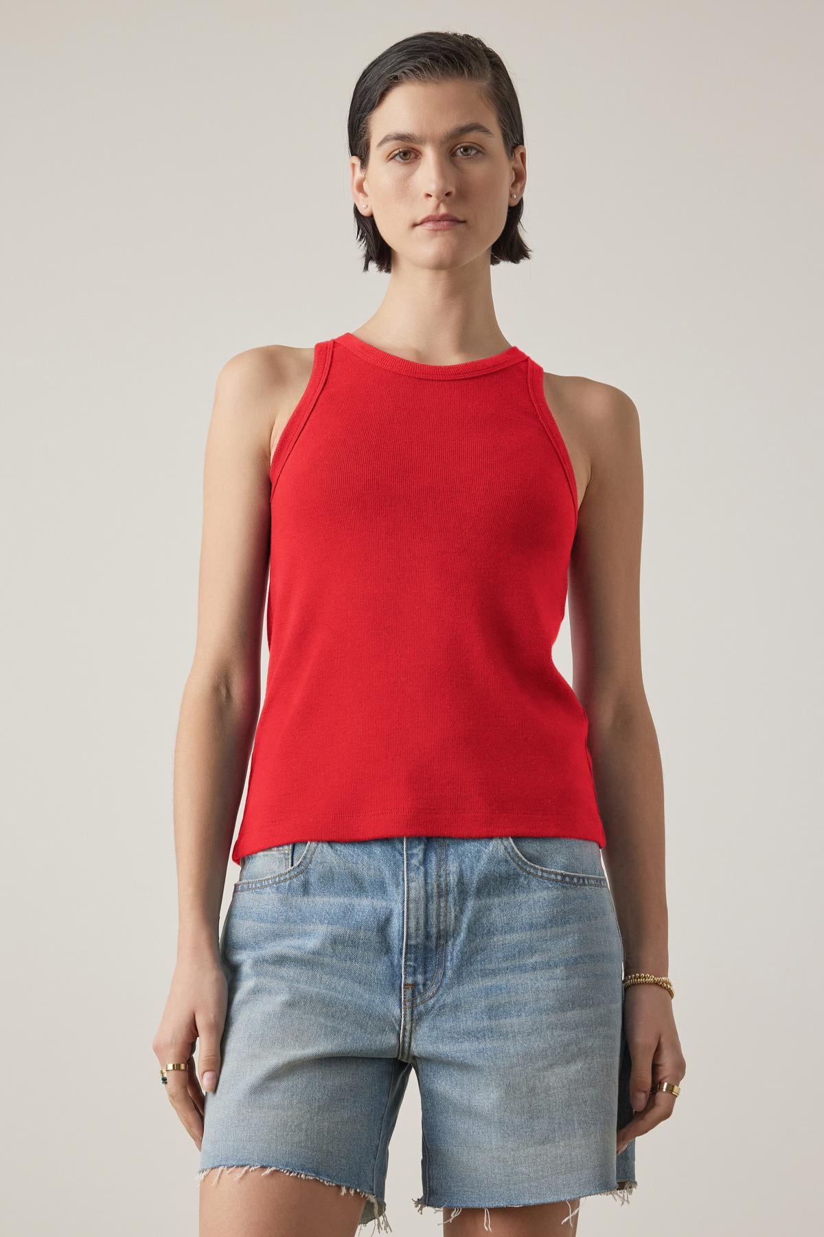   A person wearing the CRUZ TANK TOP from Velvet by Jenny Graham in a red sleeveless design, paired with denim shorts, standing against a plain background. 