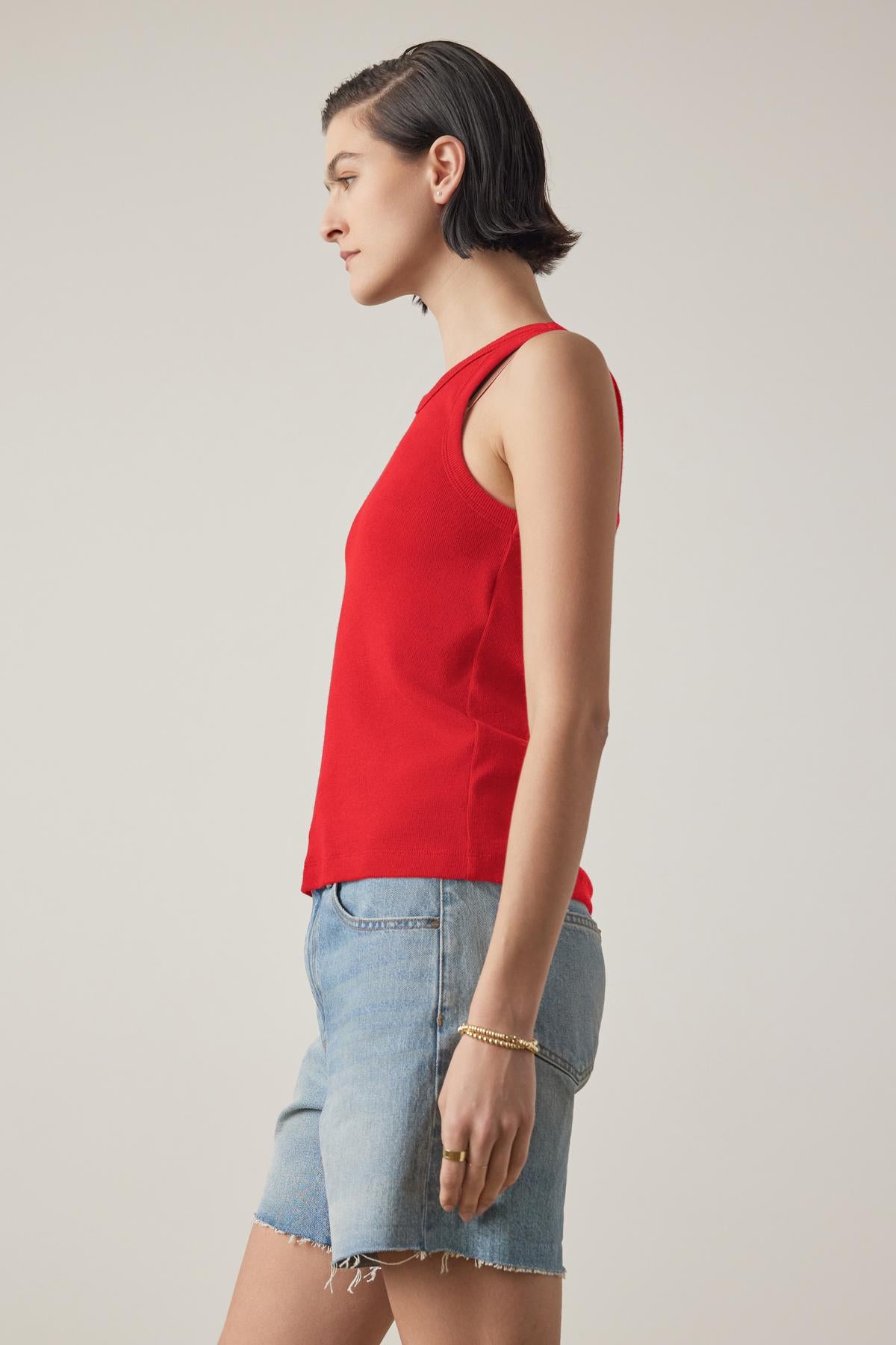 A person in the CRUZ TANK TOP by Velvet by Jenny Graham, a sleeveless red piece crafted from a ribbed cotton-modal blend, stands in profile against a plain background. This versatile top is an essential addition to any capsule wardrobe.-38333197811905