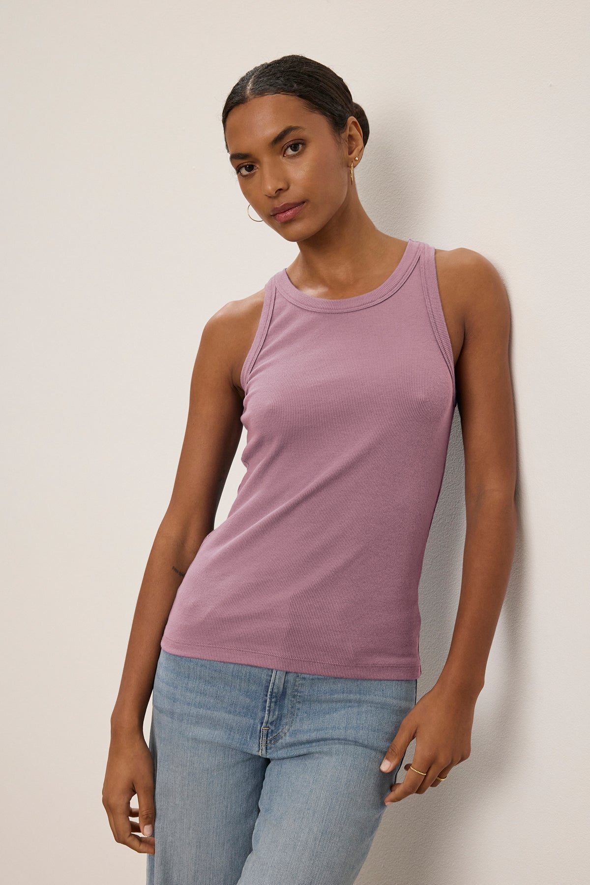   A person in the Velvet by Jenny Graham CRUZ TANK TOP, a sleeveless lavender piece made of ribbed cotton-modal for stretch, and blue jeans leans casually against the wall. 