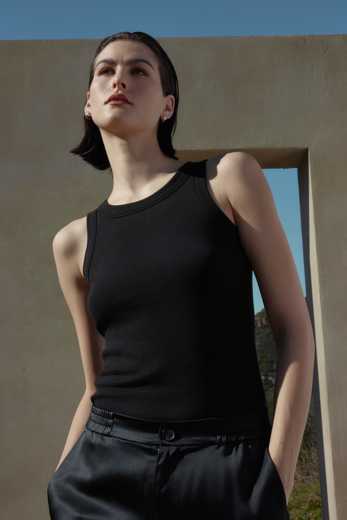   A person with shoulder-length dark hair wearing a black sleeveless top and the MODESTO SILK CHARMEUSE PANT by Velvet by Jenny Graham stands in front of a textured wall with a rectangular opening, looking upwards. 