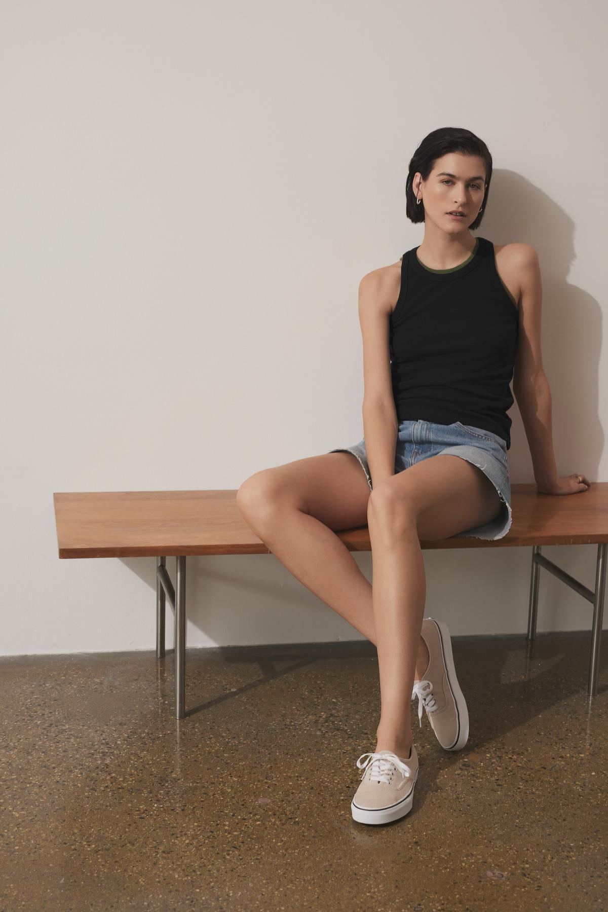 A person with short dark hair, dressed in a black CRUZ TANK TOP by Velvet by Jenny Graham, denim shorts, and white sneakers, sits on a wooden bench against a plain wall.-37676038226113