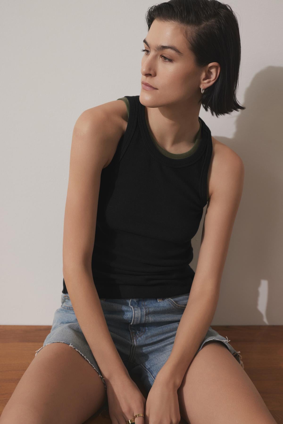   A person with short dark hair and a serious expression sits on a wooden surface, wearing the CRUZ TANK TOP by Velvet by Jenny Graham and denim shorts, looking to the side. 