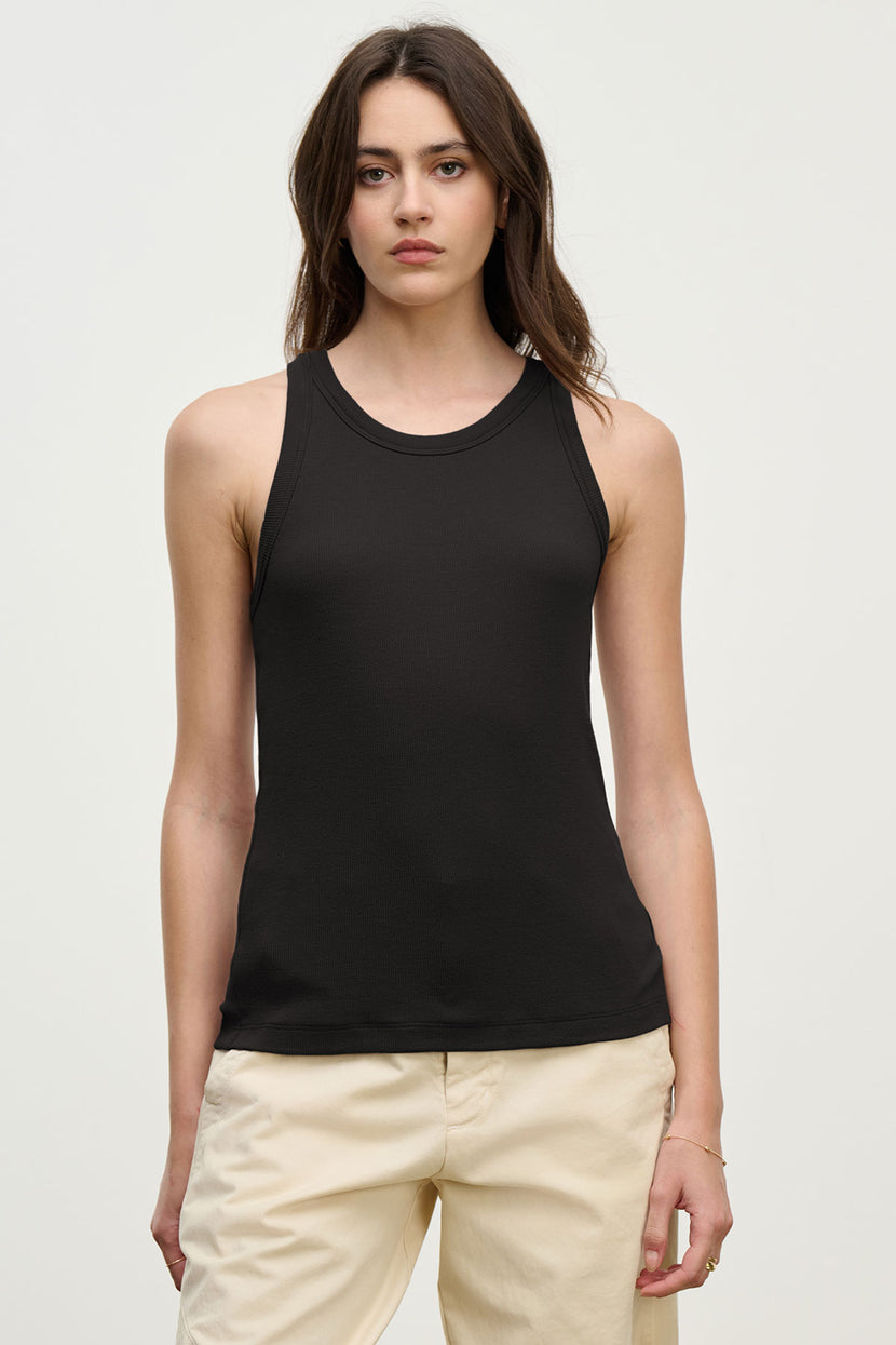 A woman with long brown hair models the Velvet by Jenny Graham CRUZ TANK TOP, a sleeveless black ribbed piece paired with light pants against a plain backdrop—a versatile staple for any capsule wardrobe.