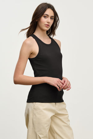 A woman wears the CRUZ TANK TOP by Velvet by Jenny Graham in black, paired with beige pants, standing against a light background—a versatile addition to any capsule wardrobe.