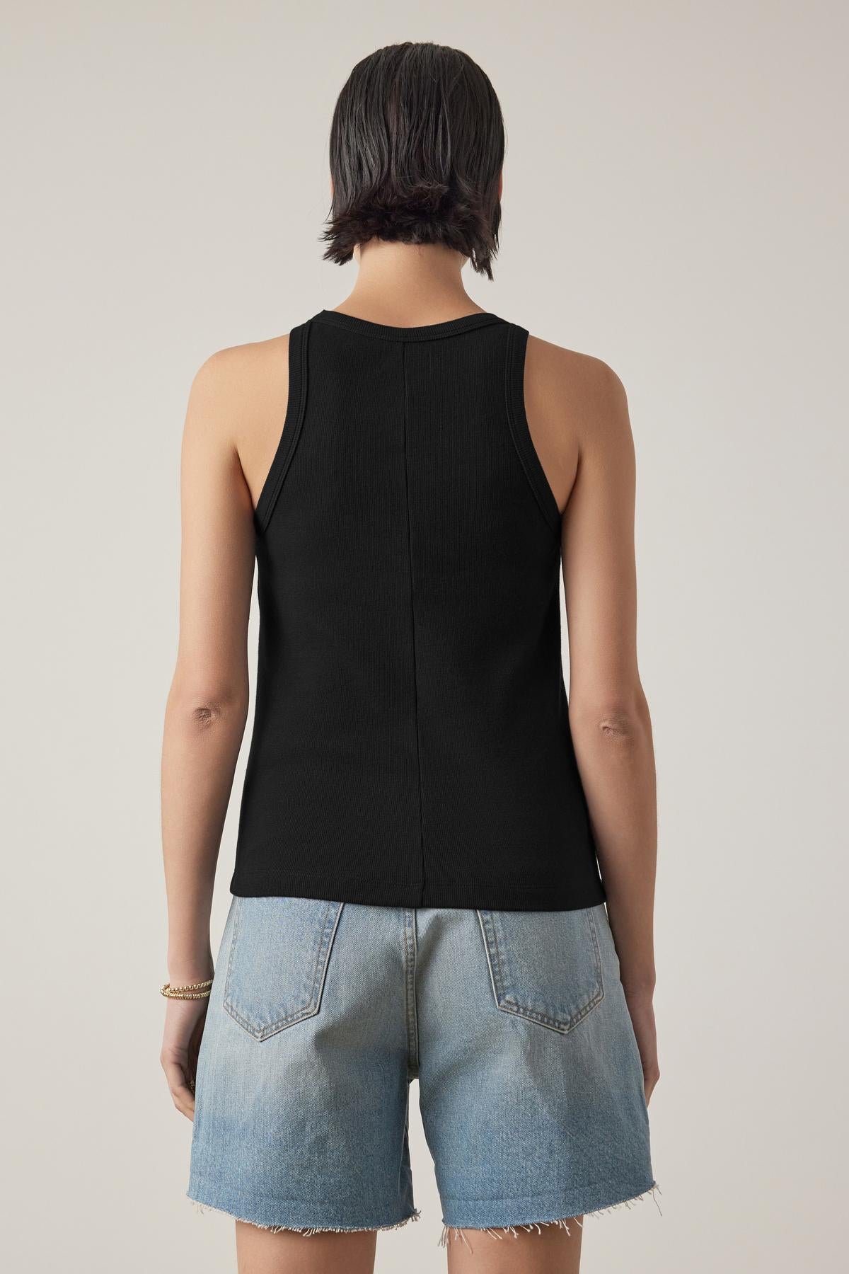   Individual in a black CRUZ TANK TOP by Velvet by Jenny Graham and denim shorts stands facing away from the camera against a plain background. 