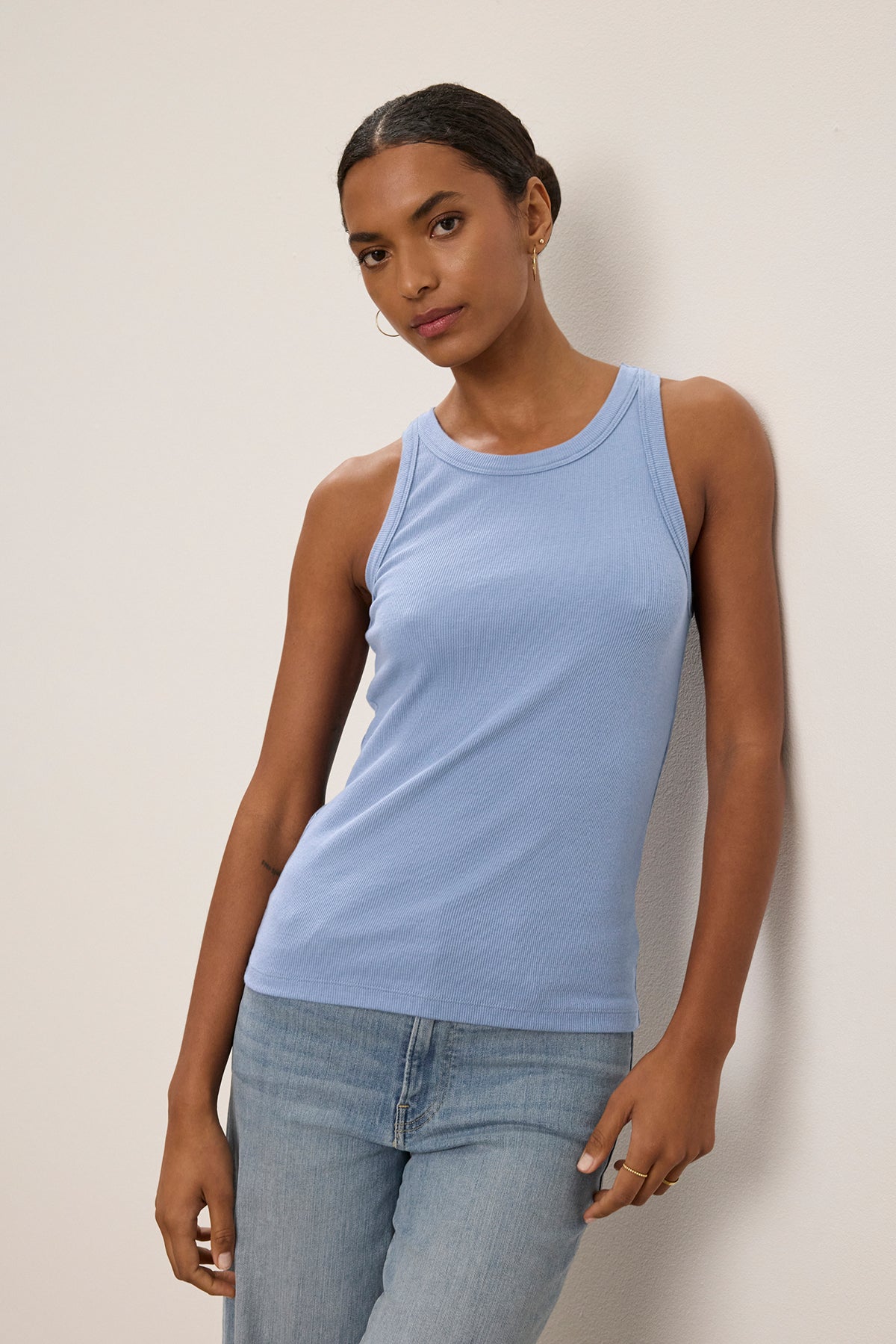   A person in a CRUZ TANK TOP by Velvet by Jenny Graham and jeans leans against a light-colored wall, looking at the camera. 