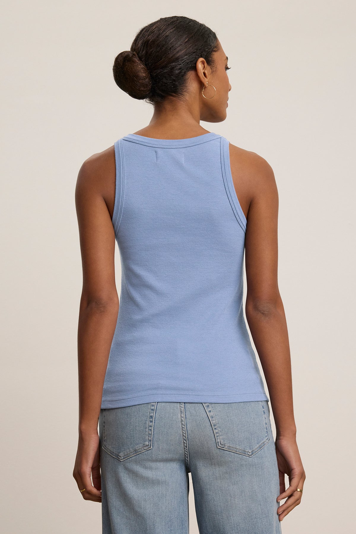   A person with a bun is seen from the back in blue jeans and the CRUZ TANK TOP by Velvet by Jenny Graham, a blue sleeveless piece crafted from a ribbed cotton-modal blend. Perfect for any capsule wardrobe, this top stands against a neutral background. 