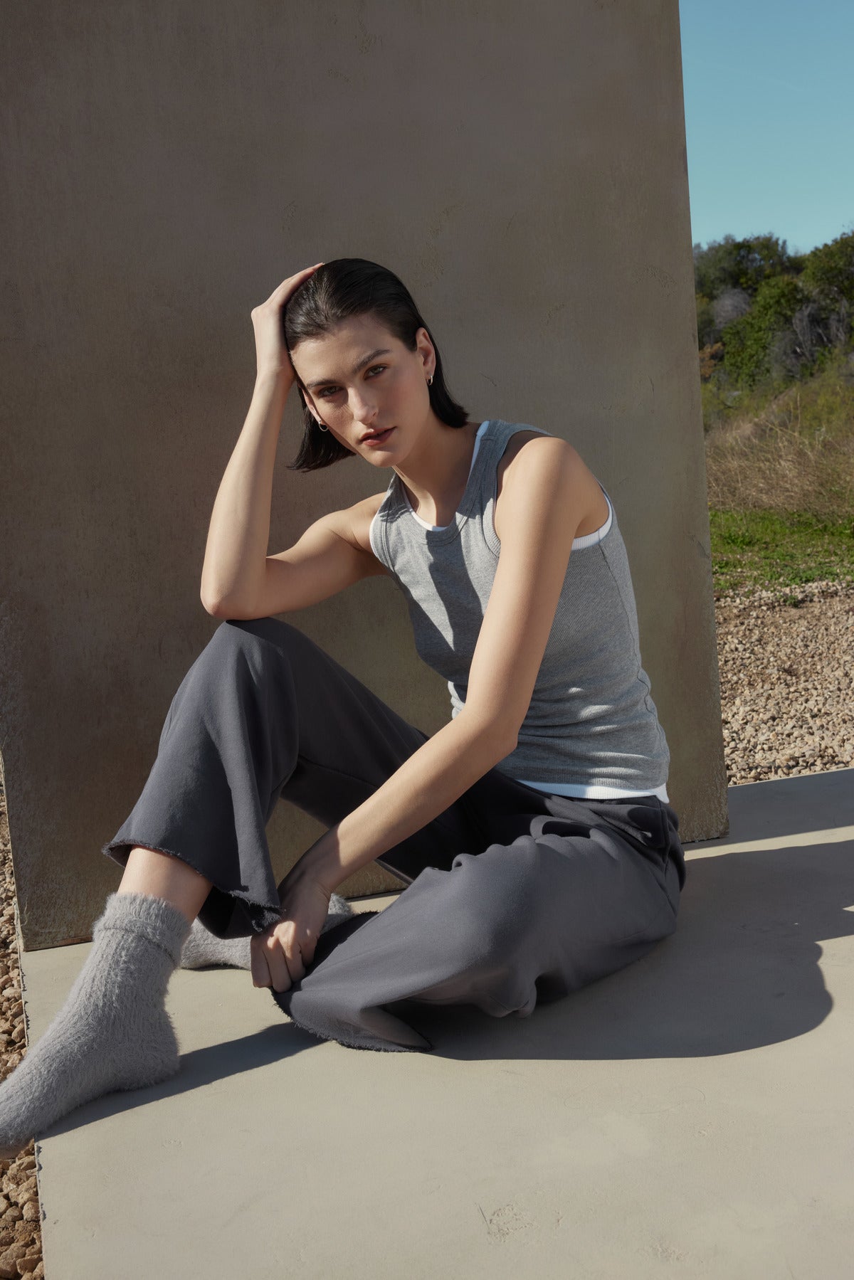 A person with short dark hair in a versatile tank and sweatpants sits on a concrete surface, resting their head on one hand, with grass and trees in the background. The ribbed cotton-modal blend of the Velvet by Jenny Graham CRUZ TANK TOP in gray adds a touch of effortless style, perfect for any capsule wardrobe.-37666964144321