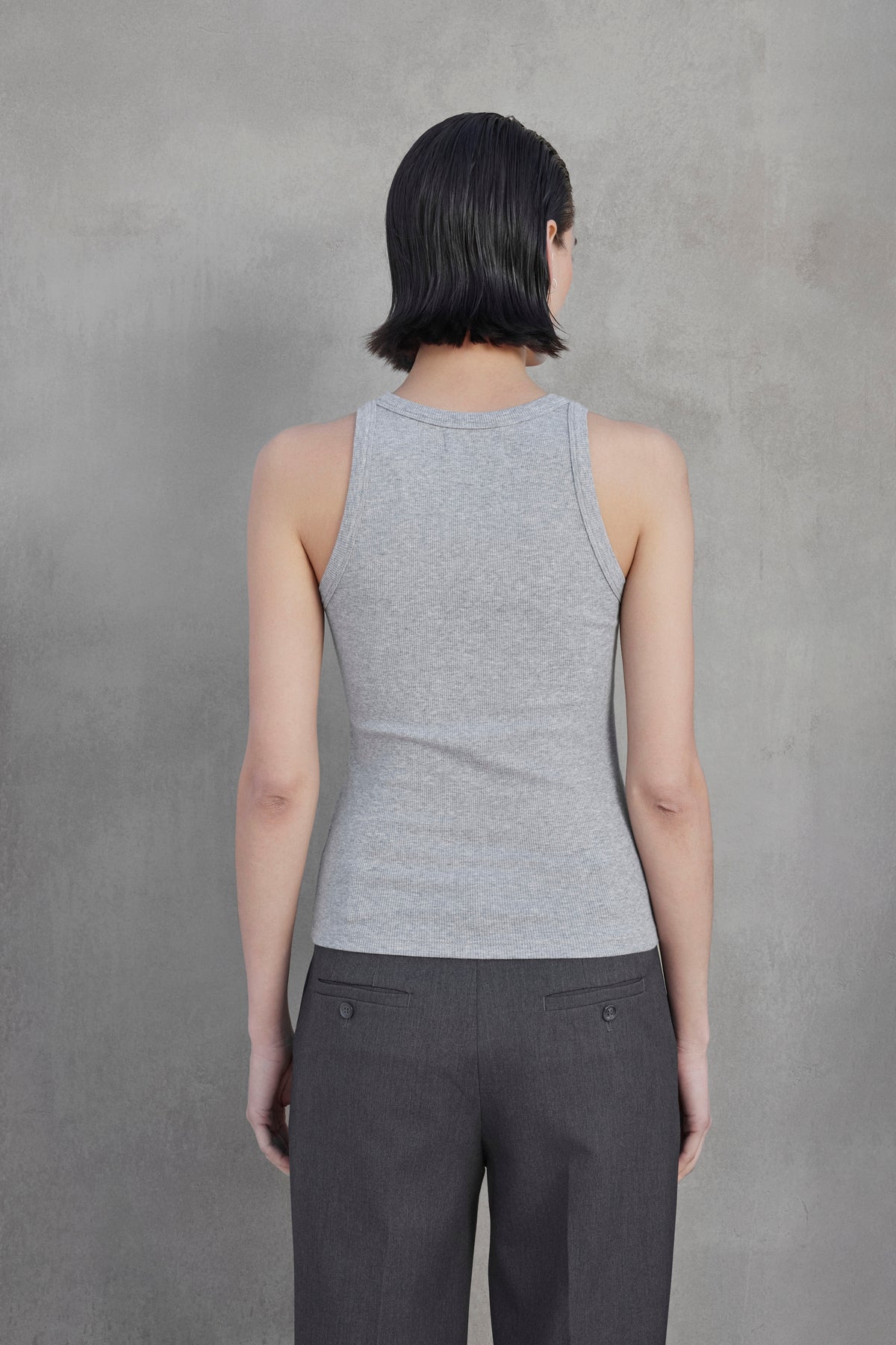   A person with short dark hair is standing facing a gray wall, wearing the CRUZ TANK TOP by Velvet by Jenny Graham, made from ribbed cotton-modal blend, and dark gray pants. Their back is to the camera. 