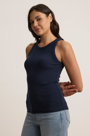 A person wearing the Velvet by Jenny Graham CRUZ TANK TOP, a sleeveless navy blue piece, paired with light jeans, stands elegantly against a neutral background—an ideal piece for any capsule wardrobe.