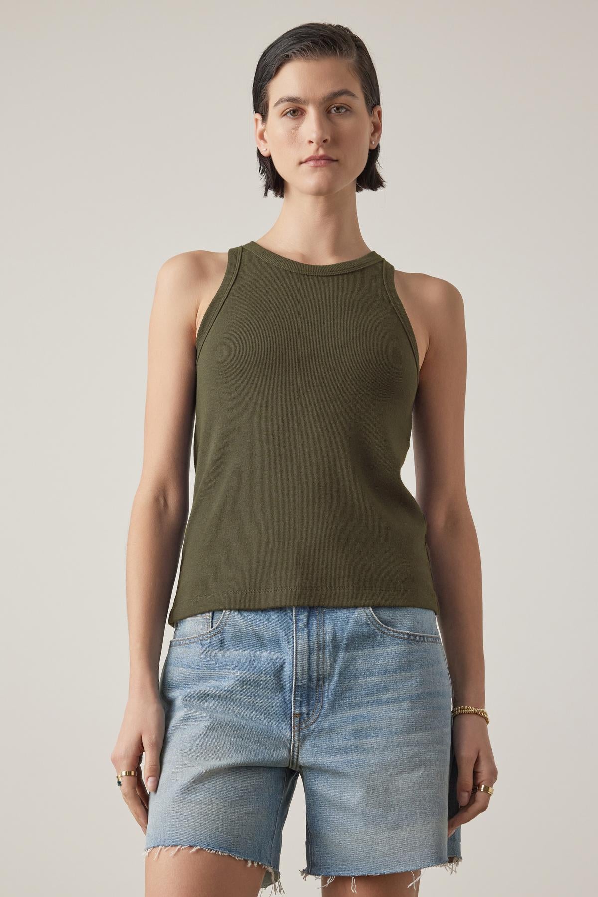   A person with short dark hair is wearing a CRUZ TANK TOP by Velvet by Jenny Graham, styled as a versatile sleeveless green top paired with denim shorts, standing against a plain background. 