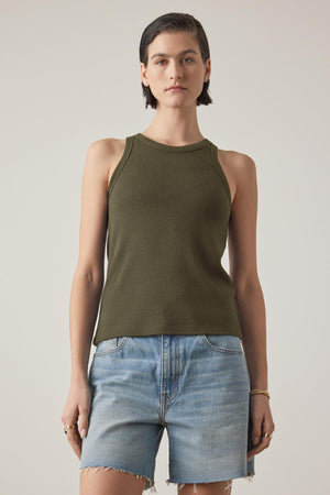 A person with short dark hair is wearing a CRUZ TANK TOP by Velvet by Jenny Graham, styled as a versatile sleeveless green top paired with denim shorts, standing against a plain background.