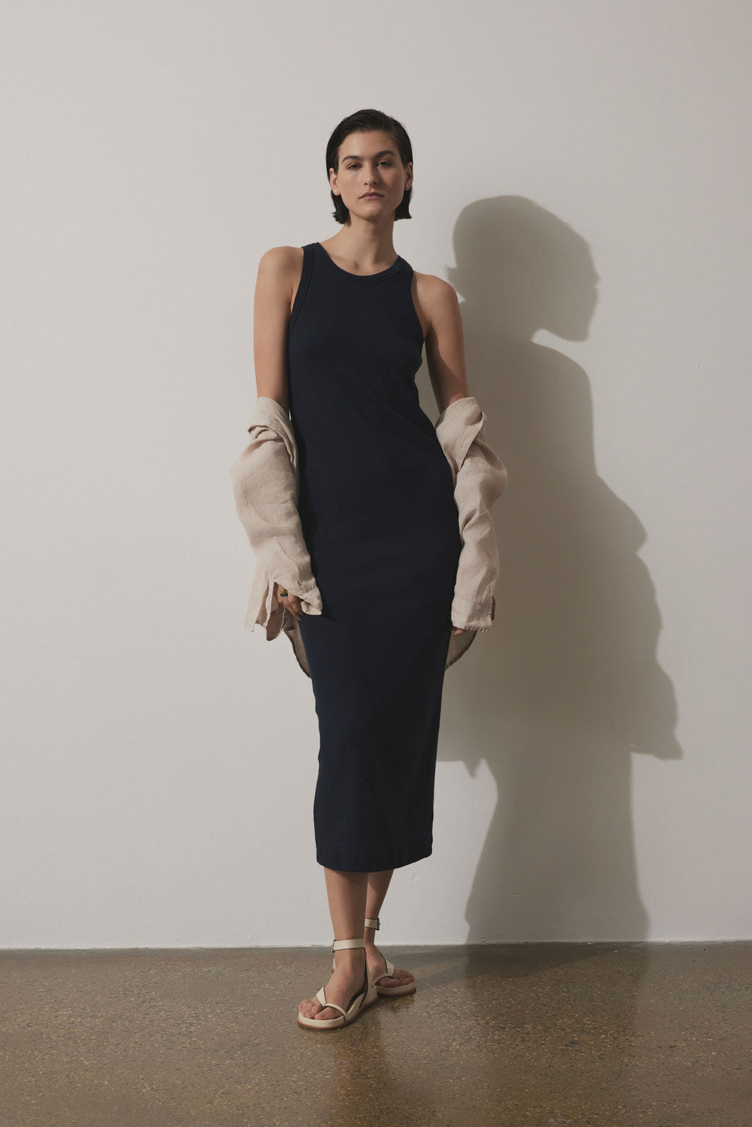   A woman in a GRIFFITH DRESS by Velvet by Jenny Graham with a crew neckline stands holding a cream jacket, with her shadow visible on the wall behind her. She wears beige sandals. 