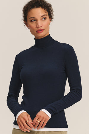 A woman wears the LINDEN TEE by Velvet by Graham & Spencer in navy blue stretch modal rib, layered over a white shirt for a chic double-layered look against a plain light background.