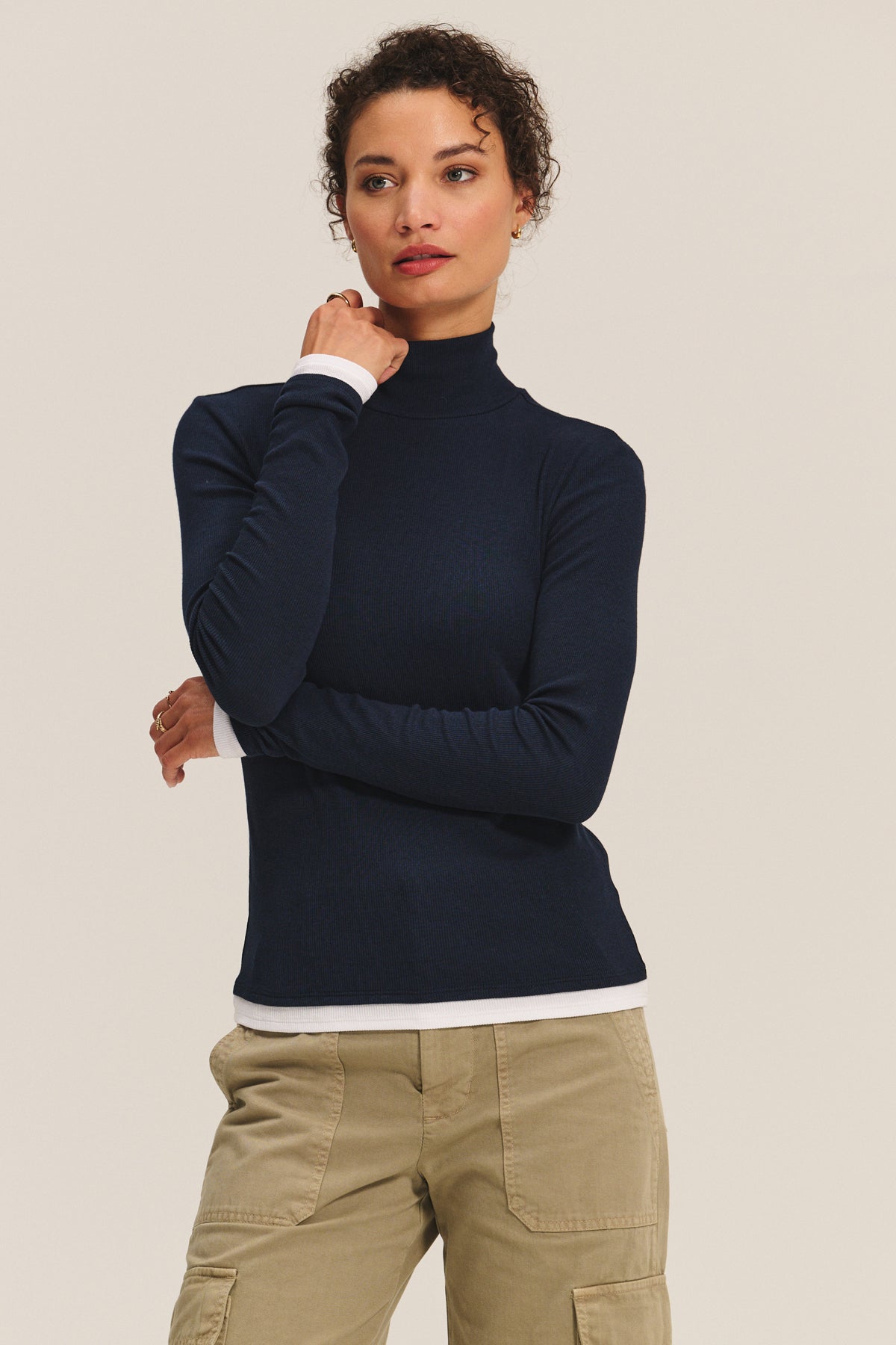 A woman poses against a plain background, wearing the LINDEN TEE by Velvet by Graham & Spencer in navy over a white shirt with khaki cargo pants for a chic, double-layered style, her hand resting thoughtfully on her chin.