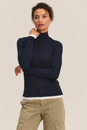 A woman poses against a plain background, wearing the LINDEN TEE by Velvet by Graham & Spencer in navy over a white shirt with khaki cargo pants for a chic, double-layered style, her hand resting thoughtfully on her chin.