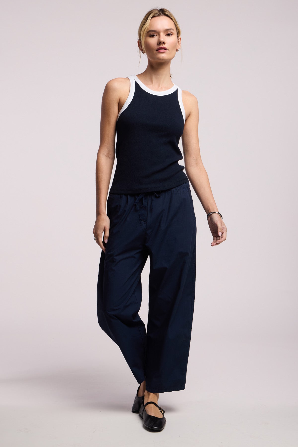 A person wears the LINDY TANK TOP by Velvet by Graham & Spencer—a black and white ribbed, fitted tank—paired with navy wide-leg pants and black shoes, standing against a plain light background.