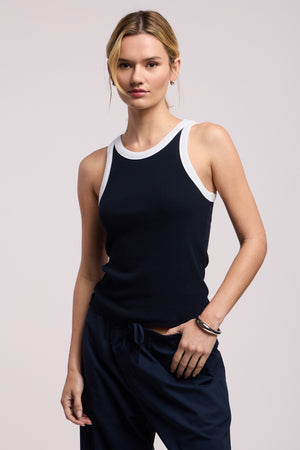 A woman with blonde hair wears the Velvet by Graham & Spencer LINDY TANK TOP, a fitted black ribbed tank with white trim, paired with dark pants, standing against a plain light background.