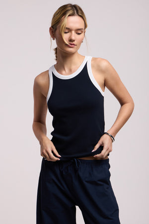 Wearing the LINDY TANK TOP by Velvet by Graham & Spencer, featuring a black ribbed design with white trim, paired with black pants. The fitted silhouette offers a casual yet modern style against a plain light background.