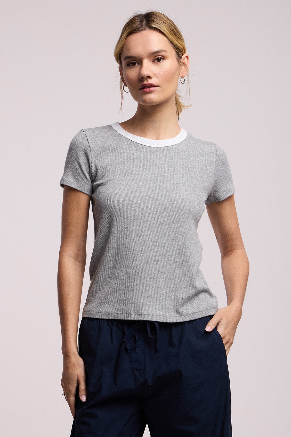 A woman stands against a plain background wearing the Velvet by Graham & Spencer MILEY TEE in light grey with a white crew neckline, paired with dark navy pants.