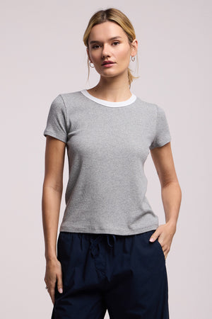 A woman stands against a plain background wearing the Velvet by Graham & Spencer MILEY TEE in light grey with a white crew neckline, paired with dark navy pants.