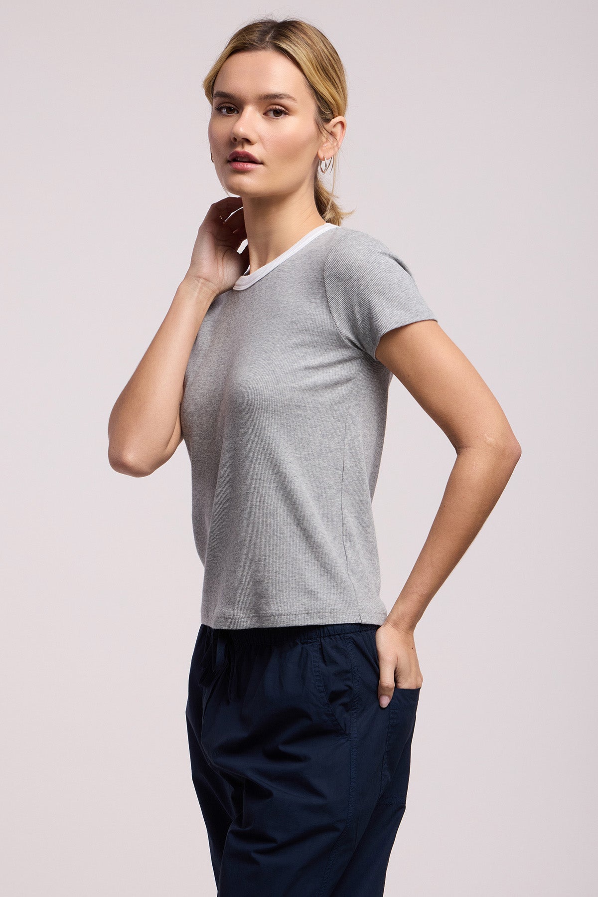 A person poses against a plain background, wearing the MILEY TEE by Velvet by Graham & Spencer, a grey crew neck short-sleeve t-shirt, and dark blue pants, with one hand on their neck and the other in their pocket.-41588377321665