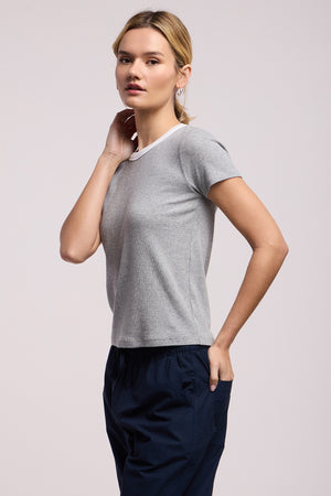 A person poses against a plain background, wearing the MILEY TEE by Velvet by Graham & Spencer, a grey crew neck short-sleeve t-shirt, and dark blue pants, with one hand on their neck and the other in their pocket.